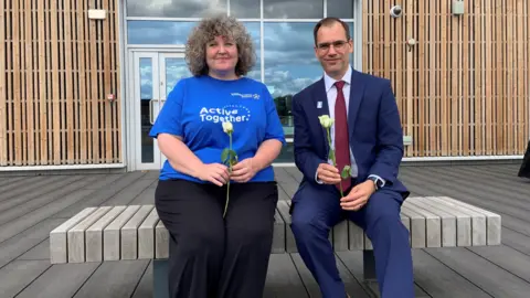 A woman with grey curly hair is sitting outdoors on a bench and smiling. She is wearing a bright blue t-shirt with the words Active Together and is holding a white rose. A man is sat next to her, also holding a rose. He has black hair, glasses, a blue suit, white shirt and maroon tie