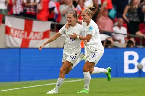 PA Media Two football players, Ella Toone and Rachel Daly, dressed in the white England strip, run towards the stands to celebrate with the fans after Toone scored at the Women's Euros in 2022. They look both happy and overcome with emotion.