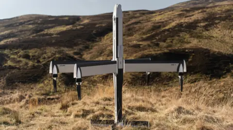 STARK A four-winged drone plane, sitting at the bottom of a grassy hill. It is white and black and standing upright