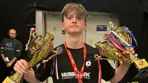 Family handout A teenage boy stands wearing a black t-shirt and holding three gold trophies and with a medal around his neck.