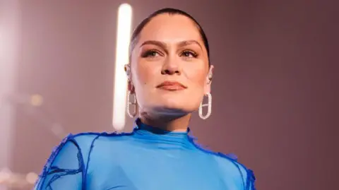 Getty Images Jessie J wearing a blue lycra stage costume and large diamond earrings, smiling slightly on stage