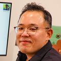 Young Hwan Chang, Ph.D., has short, wavy dark hair, eye glasses, and a black shirt, sitting at his desk with a computer screen showing data.