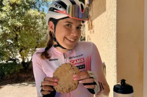 Amy Cycling Adventures Mrs Hudson wearing pink and white cycling gear, sitting down eating a biscuit. There is a water bottle on the table in front of her.