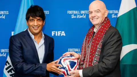 FIFA Syed Mohsin Gilani is on the left in a blue shirt and navy blazer. Infantino is on the right with a suit on and red shawl around his neck. Both of them are holding a football which is in the middle of them. It has blue and red wavy lines going through. In the back is a board with the word FIFA written on it.