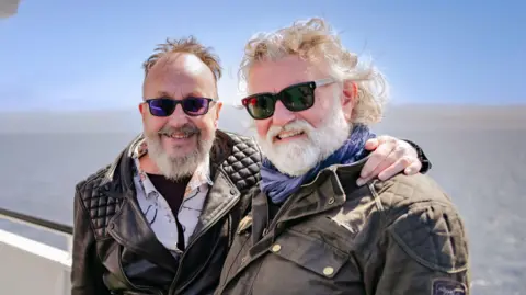 BBC / South Shore Productions Hairy Bikers Dave Myers and Si King are standing on a boat out in the water and smiling at the camera. Myers, on the left, has his arm around King and is wearing a black leather jacket. King is wearing a brown jacket and they are both wearing sunglasses.