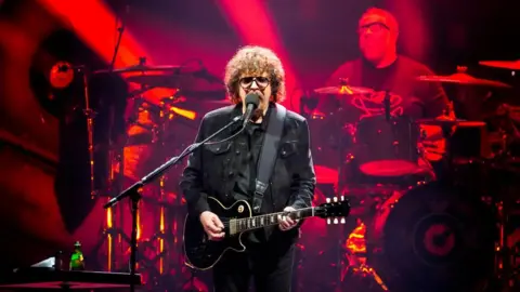 Getty Images A man with long curly brown hair in dark clothing, holding a black guitar in front of a band lit with red lights