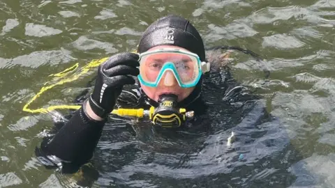 Ant Crocker A person in a wetsuit and diving gear, partially submerged in water, holding a small ring close to their face.