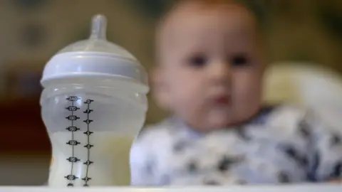 PA Media Bottle filled half-filled with baby milk formula with baby sat in a high chair out of focus in the background.