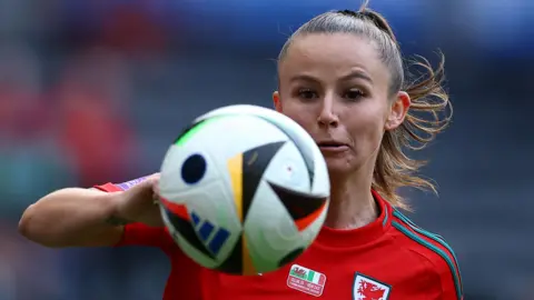 Reuters Hannah Cain shown behind a football which is photographed in the air in front of her. Her right arm is folded behind the ball at the left of the picture. She is wearing red Welsh football kit.