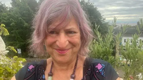 Judith Levy A woman with pink and grey hair lookings into the camera, half-smiling. She wears a beaded necklace and a colourful shirt. Behind her there is some vegetation.