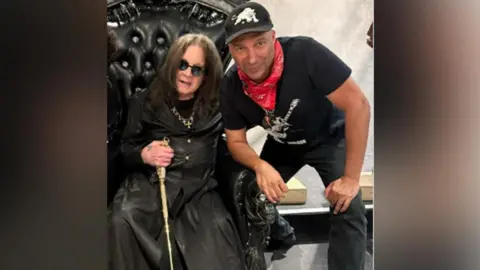 TOM MORELLO/INSTAGRAM Ozzy Osbourne dressed in long black leather, sat in a black throne with his arm around Tom Morello, wearing a black t shirt and trousers, a black cap and red bandana
