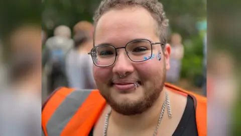 Nathan Maradei wearing a black shirt underneath an orange hi-vis vest, and black square glasses. He has a silver chain around his neck, a septum piercing, a several rings on his lower lip. He is smiling at the camera, with blue glitter dotted on his cheekbones.