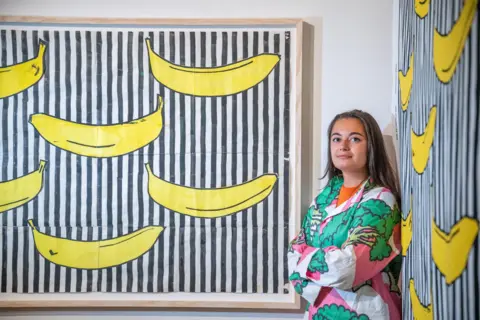 Phil Wilkinson A young woman stands wearing one of the broccoli shirts as she leans against the Randig Banan patterns on two walls - it is a bold black and white striped pattern with large, cartoon bright yellow bananas