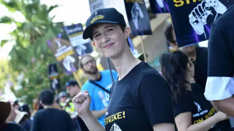 Getty Images A woman with short black hair wears a black baseball cap and t-shirt with matching Video Game Actors' strike logos, with text and a picture of a hand holding a control pad.