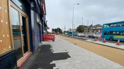 Ed Barnes A paved partially paved area outside shops which unfinished roadworks and cones liming the street. Traffic is queuing on the main road.