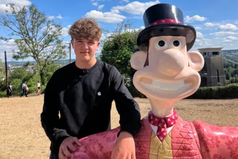 Barney, now 15, stands next to a life-size sculpture of Wallace. He is wearing a black jumper and has one arm casually resting across Wallace’s shoulder. Wallace is dressed in his classic outfit, including a red cardigan and white shirt.