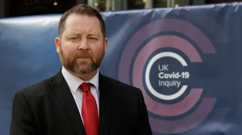 Liam McBurney/PA Wire Eddie Lynch, the former commissioner for older people for Northern Ireland, pictured during an earlier hearing in the UK Covid-19 Inquiry last year.  He has short, reddish brown hair and a greying beard.  He is wearing a black suit, a white shirt and a red tie.  He is standing against a navy blue banner which shows the inquiry's pink and white logo. 