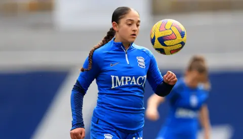 Getty Images Layla in her Birmingham City football kit which is blue. She's looking at yellow colour ball that is head height. In the background you can see her fellow footballers. 