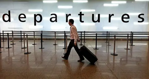 Getty Images A man wearing black trousers and a white shirt pulling a suitcase in front of a giant sign saying departures that is brightly lit from behind