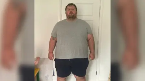 James Baker James Baker - a 38st man with brown hair and facial hair - before losing half his weight. He is wearing a grey T-shirt and black shorts.