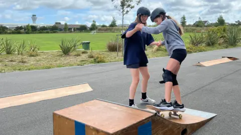 BBC Two individuals are on a paved path in an outdoor setting with greenery and cloudy skies. One person is standing on a skateboard at the top of a small wooden ramp, while the other is assisting by holding their hands. Both are wearing helmets, and one is also wearing knee pads