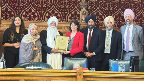 Nishkam Media A group of MP's standing next to Bhai Sahib Ji who is wearing a white turban, black coat and white cloth around his neck. He holds and award being handed to him by Seema Malhotra MP who wears a red jacket and dotted top.