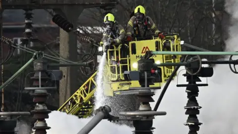 Getty Images Two fire fighters wearing breathing apparatus are on board a platform which has lifted them above the electrical substation which caught fire in March. One is holding a hose which is dampening down the flames. Smoke is billowing.