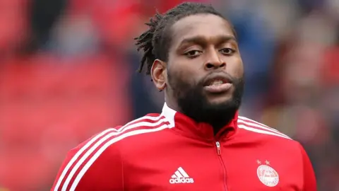 PA Media Jay Emmanuel-Thomas wearing a red Aberdeen tracksuit top with white stripes. A crowd is blurred in the background.