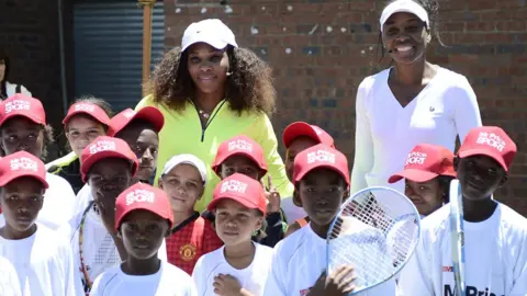AFP/Getty Images US tennis player Serena Williams (left) in a yellow top and white cap flanked by her sister Venus in a white top and cap pose with children in red caps and white T-shirts, some holding rackets after a two-hour tennis clinic at the Arthur Ashe Soweto Tennis Centre - November 2012.