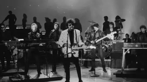 Getty Images A black and white image of Jeff Lynne fronting the original line-up of the Electric Light Orchestra, playing live in 1972. He has his trademark sunglasses on and is wearing a light coloured suit jacket and black trousers, and is holding an electric guitar.