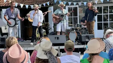 Dementia Friendly Guernsey Four men performing as part of a band on stage at an outdoor event. In the crowd are men and women wearing hats.