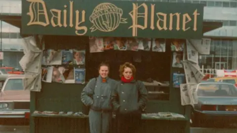 Stephanie English Two people standing in front of a green "Daily Planet" newsstand with newspapers and magazines. Stephanie English is on the right. Parked cars and buildings are in the background.