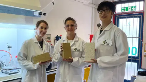 Tom MacDougall/BBC Three members of the earth4Earth company, wearing white lab coats, in their research facility at Sheffield Technology Parks. They each hold one of the "carbon absorbing" bricks. Two of the bricks are beige in colour and the other brick is an off-white colour.