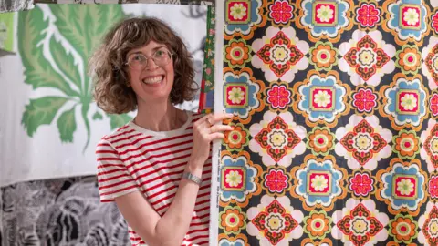 Phil Wilkinson Anna Sandberg Falk, a smiley woman with brown curly hair, peeks out from behind a colourful tile-patterned panel of fabric - several different bold designs hang behind her.
