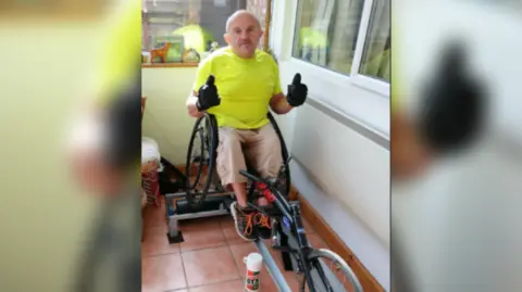 Les Hampton Les Hampton in his sports wheelchair in his conservatory at home, wearing a bright yellow t shirt and sand colour three quarter length cargo trousers. He has trainers on and black gloves, with both hands in a thumbs up sign to the camera.