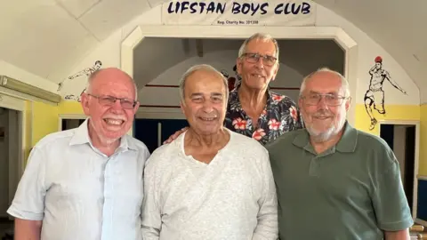 Stuart Woodward/BBC Four former members of the Lifstan Boys Club stand in front of a stage which bears the club's name. There is a domed white painted roof curving over the stage. The men are in their late 70s and early 80s. One on the left wears a light blue shirt and has a white goatee beard. Second from left is a man wearing a white open necked shirt. At the back is a taller man wearing a dark floral pattern shirt and glasses, and on the right end is a man wearing a dark green polo shirt, glasses and has a grey beard. All four men are smiling at the camera