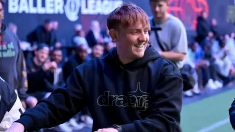 Getty Images Angryginge at a Baller League event. The streamer has red hair with a long fringe and smiles while looking over his left shoulder. He wears a black hoodie and in the background, rows of people sit pitchside. 