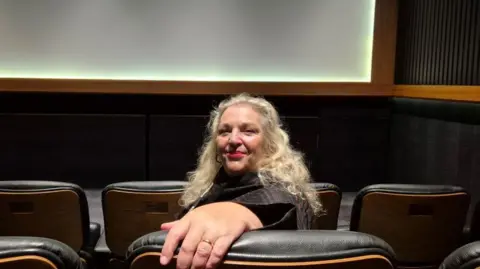 BBC Angela Freeman - a woman with long hair, sits in a cinema seat and looks to the camera - the screen is visible behind her