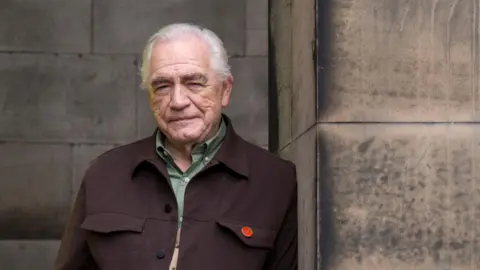 PA Media Actor Brian Cox in green shirt and brown jacket smiles as he leans against a stone plinth