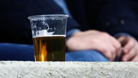 Getty Images A pint of beer in a plastic cup sitting on a wall. A person can be seen sitting in the background.