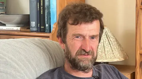 A man with a beard, wearing a grey top sitting on a sofa in his rented home.