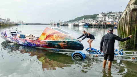 Mike Newman/ROW4MND An ocean rowing boat in bright colours being launched by a man and a woman from a trailer with docks and boats in the background.
