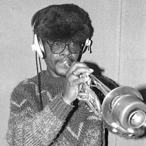 David Corio/Getty Images Black and white photo of Eddie 'Tan Tan' Thornton playing a trumpet while wearing headphones