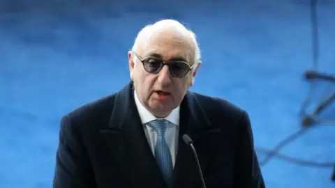 Adam Fradgley - AMA/West Bromwich Albion FC via Getty Images Barnett with thinning white hair, wearing darkened glasses, a dark overcoat, white shirt and blue tie, stands in front of a microphone in 2018.