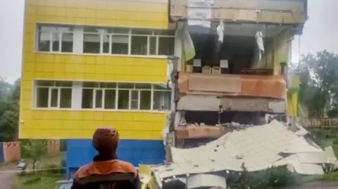 Russian Emergencies Ministry via EPA A rescue worker wearing a red hard hat stands in front of a damaged building that is two storeys tall and the front of it looks like it has been ripped away