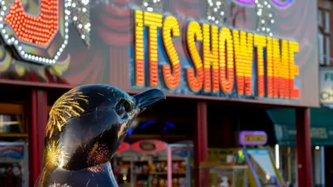 Sean Buckley A head of a penguin sculpture, which is painted black with coloured fireworks on, is at the bottom left. In the background is the outside of an arcade which says 'Its showtime' in yellow and orange.