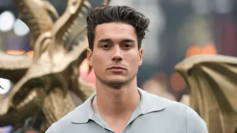 Getty Images Miles Nazaire poses in front of a Dragon statue at a Premier. He has black hair up in a cwiff. He has a blue steel pose looking into the camera. He's wearing a chain and a grey polo shirt.