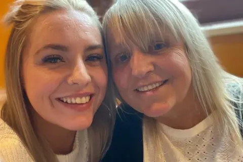 Georgina King Two women with long blonde hair, both wearing white tops, put their heads together and smile at the camera while the one on the right takes a selfie. Behind them is a yellow wall and a window with a brown frame.