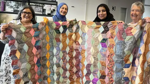 Aisha Iqbal/BBC Four ladies holding up a colourful patchwork quilt and smiling