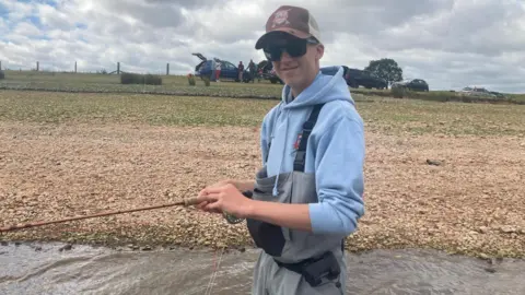 BBC A teenage boy standing in some water with a blue hoodie on and waders. He has a cap and black sunglasses and is holding a fishing rod.